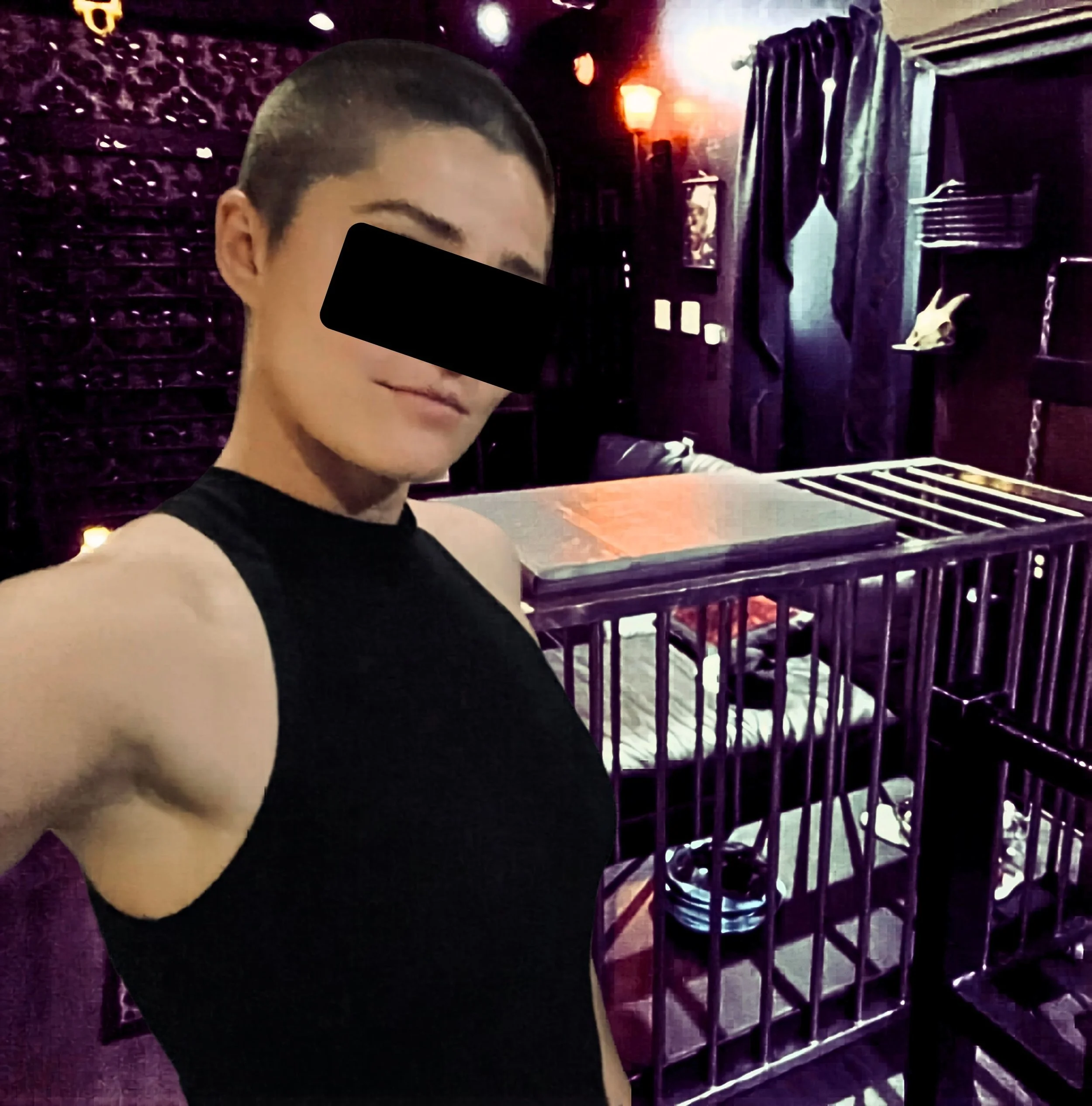 San Francisco Dominatrix in a black face mask, taking a selfie in a dimly lit purple dungeon room with a skull decoration.